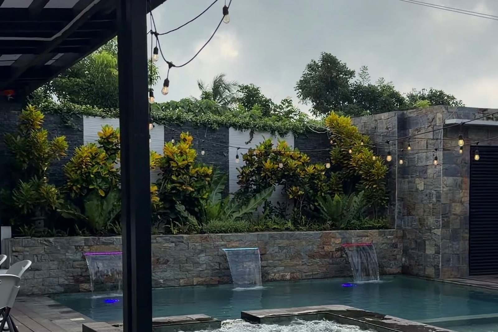 Pool with stone waterfall wall lit by string lights at dusk